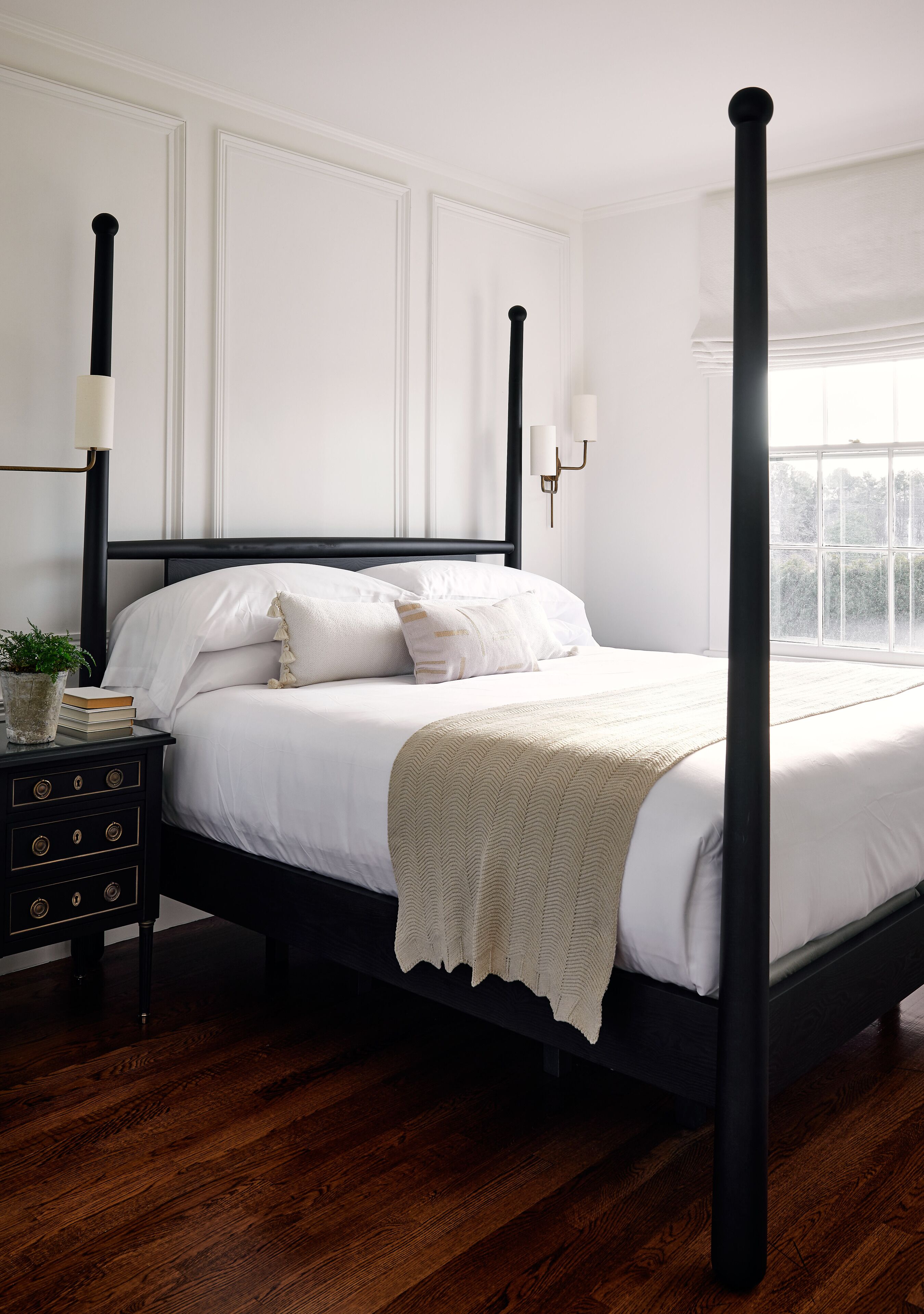 A four-poster bed with white linens, a bedside table with a plant and books, and a window with a view of greenery.