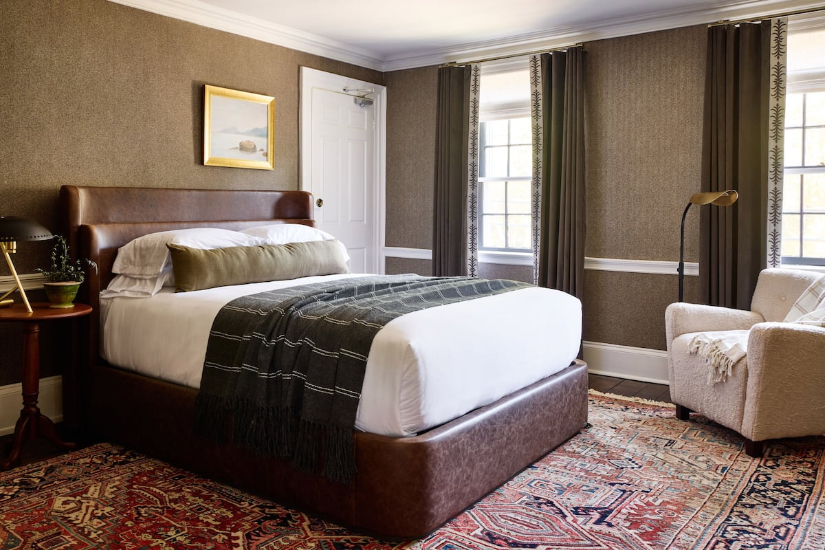 A bedroom with a large bed, a chair, a nightstand, a window with curtains, and a framed picture on the wall.