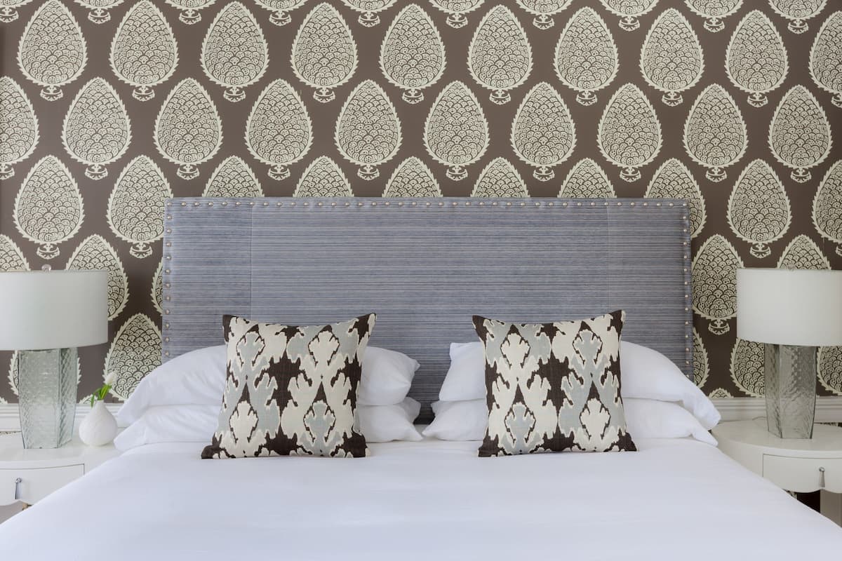 A neatly made bed with patterned pillows, a grey headboard, and a wall with a leaf pattern.
