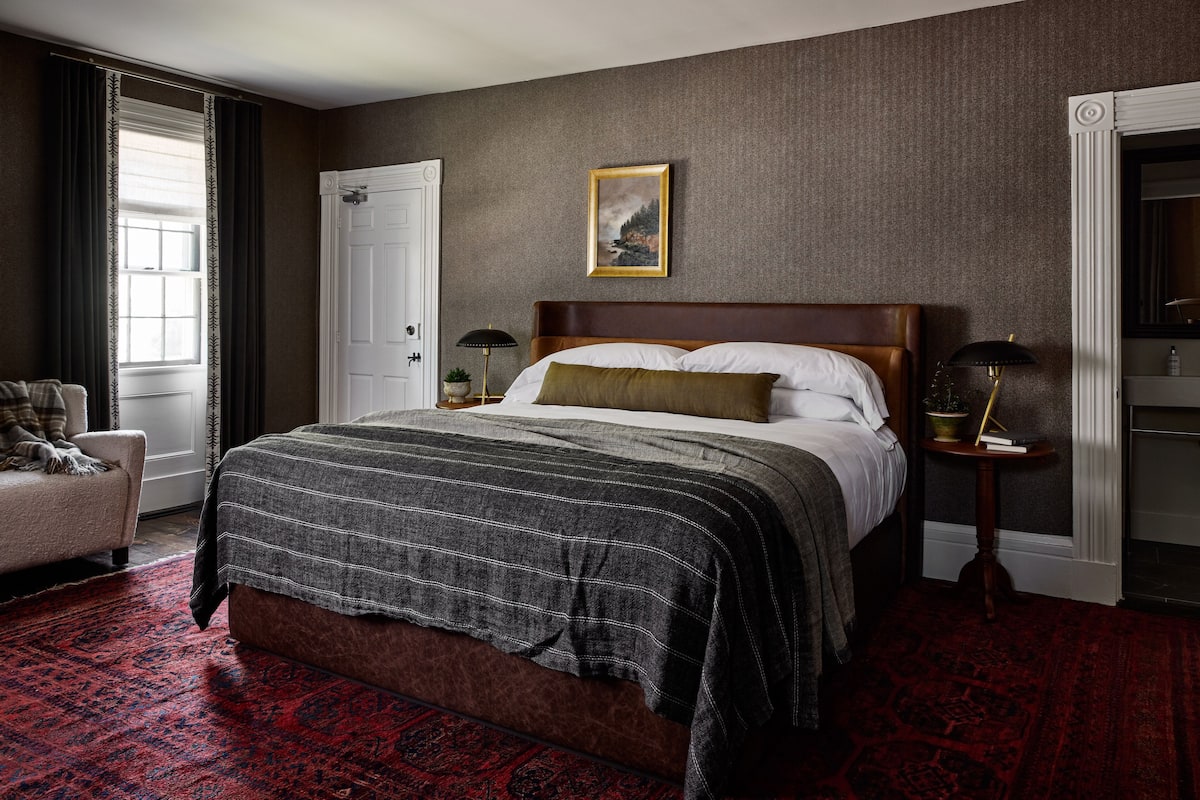 A bedroom with a large bed, a bedside table with a lamp, a door, and a window with curtains.