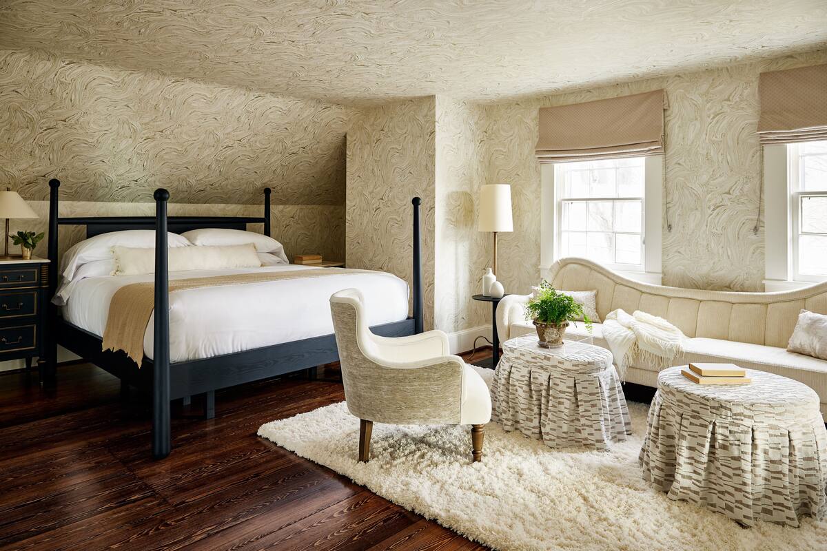 A bedroom with a four-poster bed, a sofa, two round ottomans, a lamp, and a potted plant.