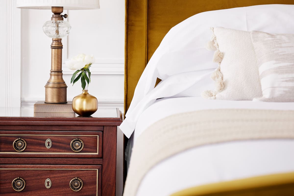 A neatly made bed with white linens and a gold headboard, a bedside lamp with a white shade, and a small vase with a single white flower on a wooden nightstand with drawers.