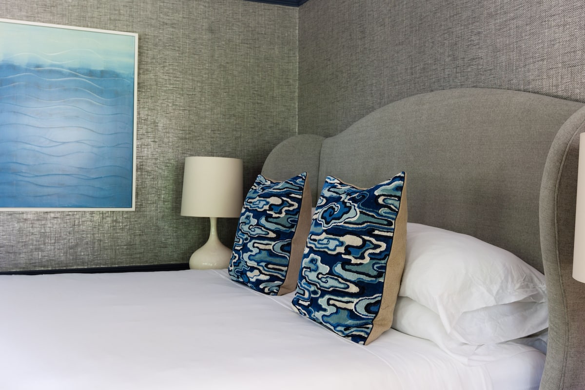 A neatly made bed with white linens, two patterned pillows, a white bedside lamp, and a large abstract painting on the wall.
