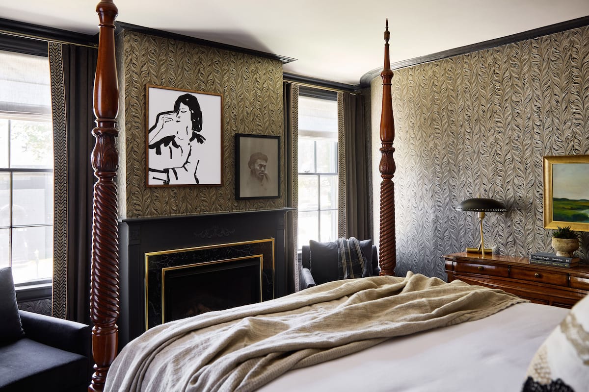 A bedroom with a four-poster bed, a fireplace, and patterned wallpaper.
