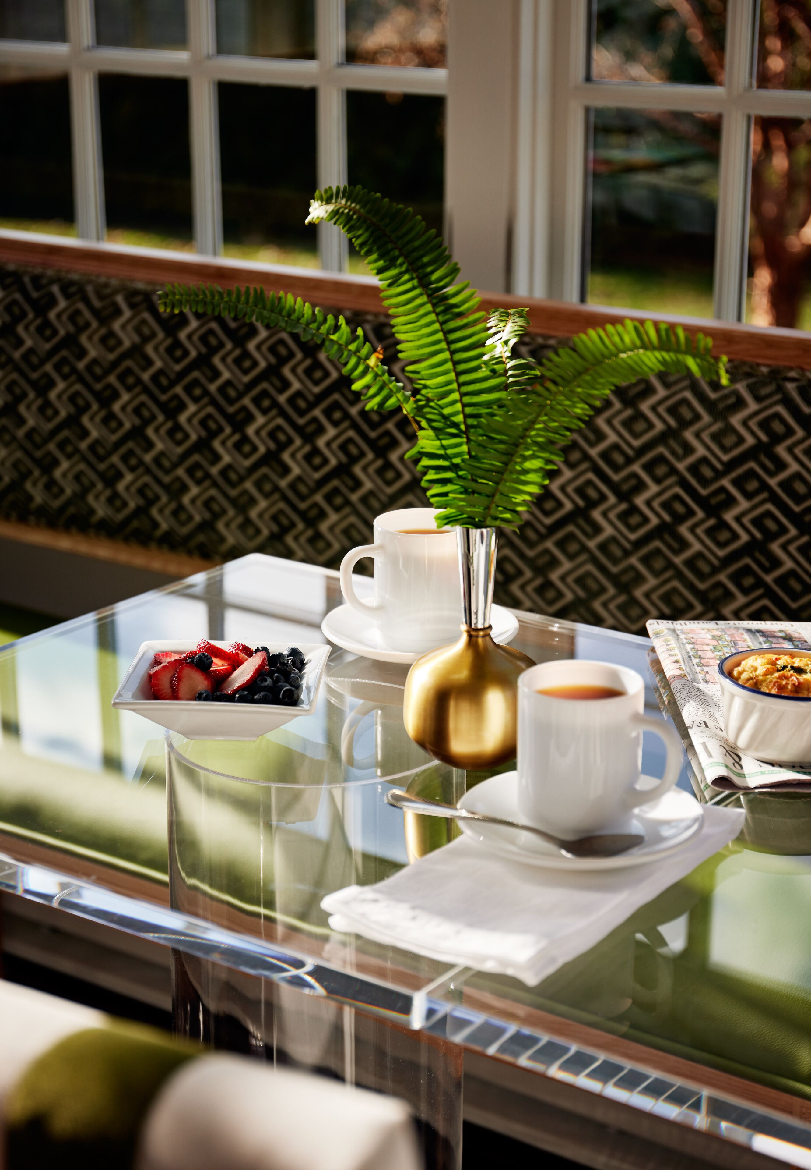 A glass table with a white cup, a gold vase with a green plant, a bowl of fruit, and a newspaper.