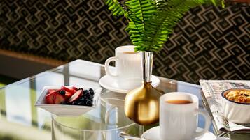 A glass table with a white cup, a gold vase with a green plant, a bowl of fruit, and a newspaper.