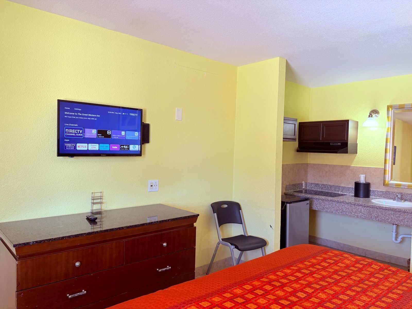 A hotel room with a bed, a desk with a computer, a chair, a small kitchenette, and a TV mounted on the wall.