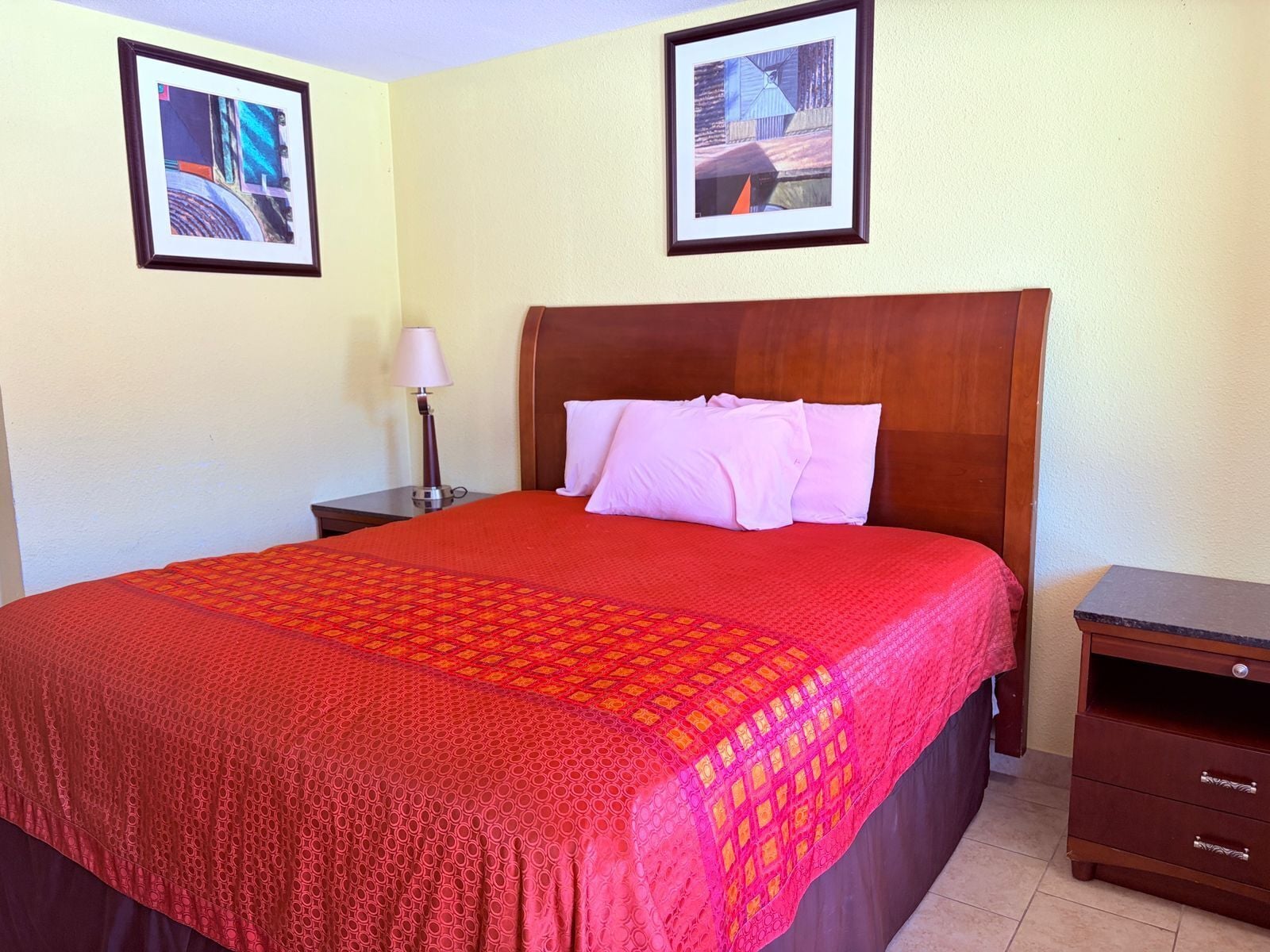 A hotel room with a large bed, a wooden headboard, two framed pictures on the wall, a nightstand with a lamp, and a dresser.