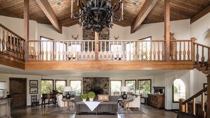 A spacious interior with a high ceiling, wooden beams, a grand chandelier, a fireplace, and a balcony with a railing.