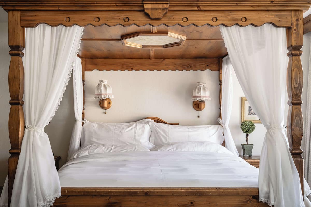 Una cama con dosel, ropa de cama blanca, cabecera de madera, dos lámparas fijadas a la pared y una pequeña planta en maceta sobre la mesita de noche.
