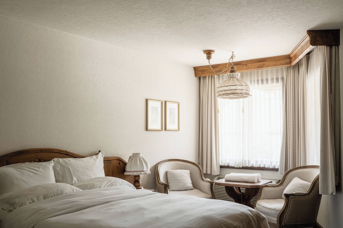 Una habitación de hotel con una cama grande, dos sillas, una mesita pequeña y una ventana con cortinas.