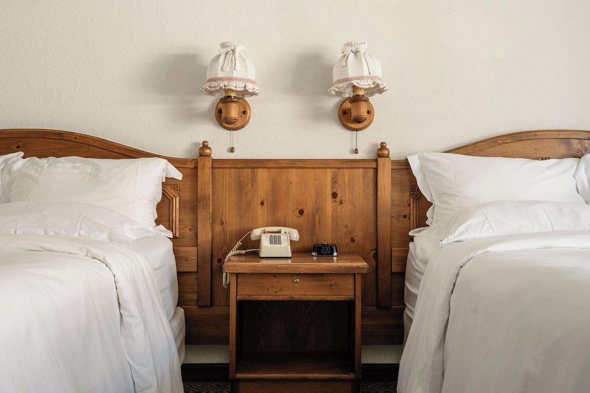 Habitación doble con dos camas de madera, una mesita de noche con teléfono y reloj, y dos lámparas de pared.