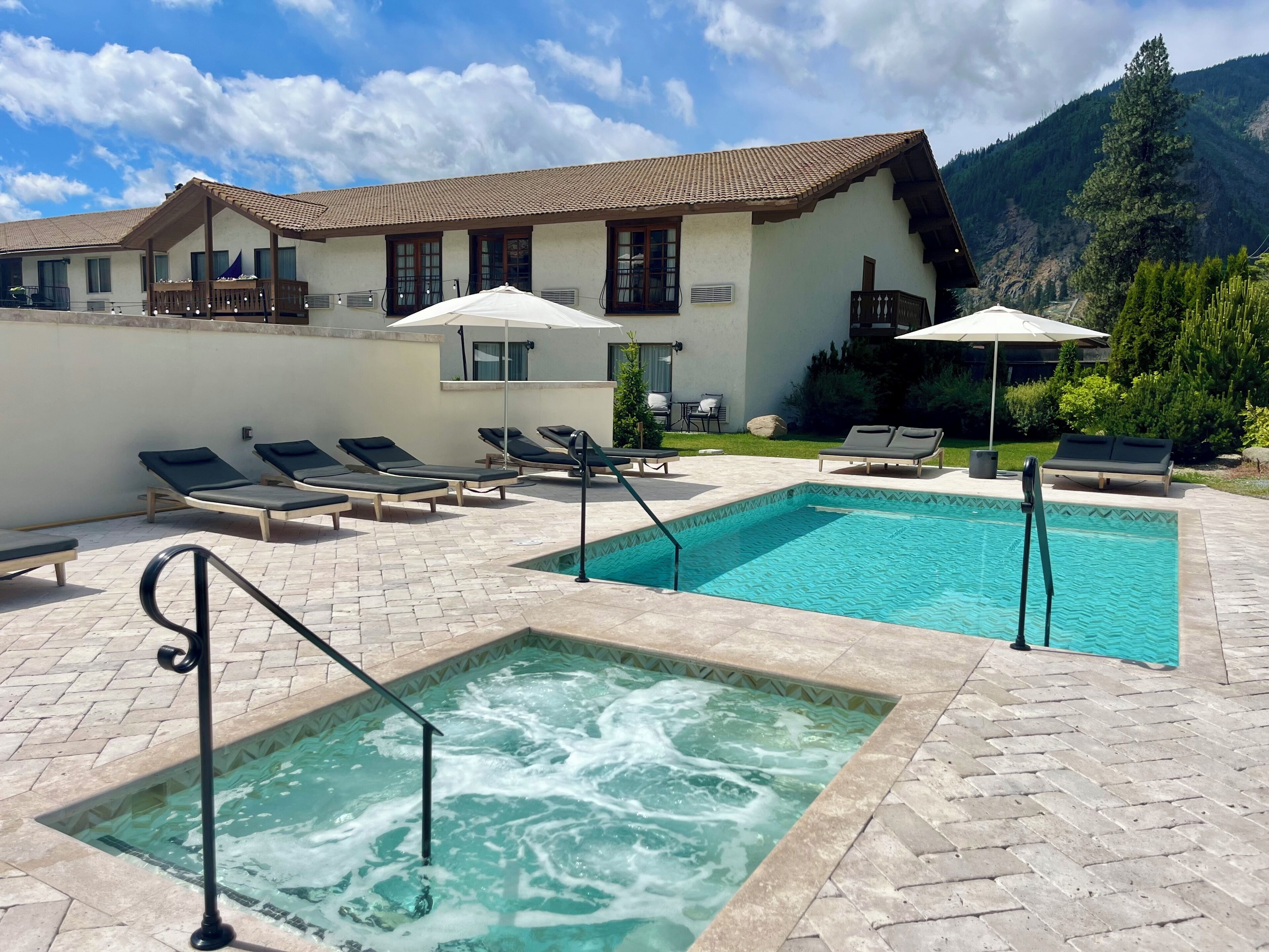 Un espace piscine doté de chaises longues et d’un jacuzzi, entouré d’un bâtiment avec un toit en tuiles et surplombant un paysage montagneux.