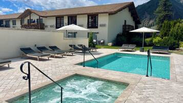 A pool area with lounge chairs and a hot tub, surrounded by a building with a tiled roof and a mountain backdrop.