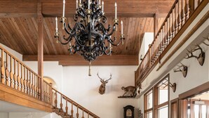 A spacious hall with a grand chandelier, wooden staircase, and deer antlers on the wall.