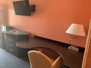 A hotel room with a desk, a chair, a television mounted on the wall, and a lamp.
