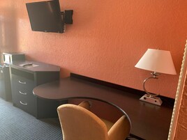 A hotel room with a desk, a chair, a television mounted on the wall, and a lamp.