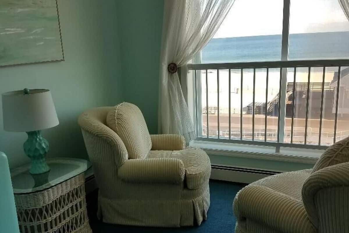 A room with a view of the sea, featuring two beige armchairs, a small round table with a lamp, and a wicker side table.