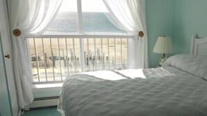 A bedroom with a bed, a window with curtains, and a nightstand with a lamp. - Mayfair Hotel (Belmar)