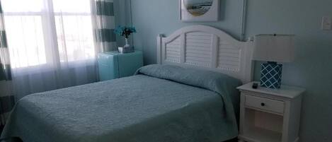 A bedroom with a bed, a nightstand, a lamp, a window with curtains, and a wall-mounted picture.
