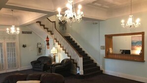 A spacious living room with a dark carpet, leather sofas, a chandelier, a staircase, and a large mirror reflecting a cityscape. - Mayfair Hotel (Belmar)