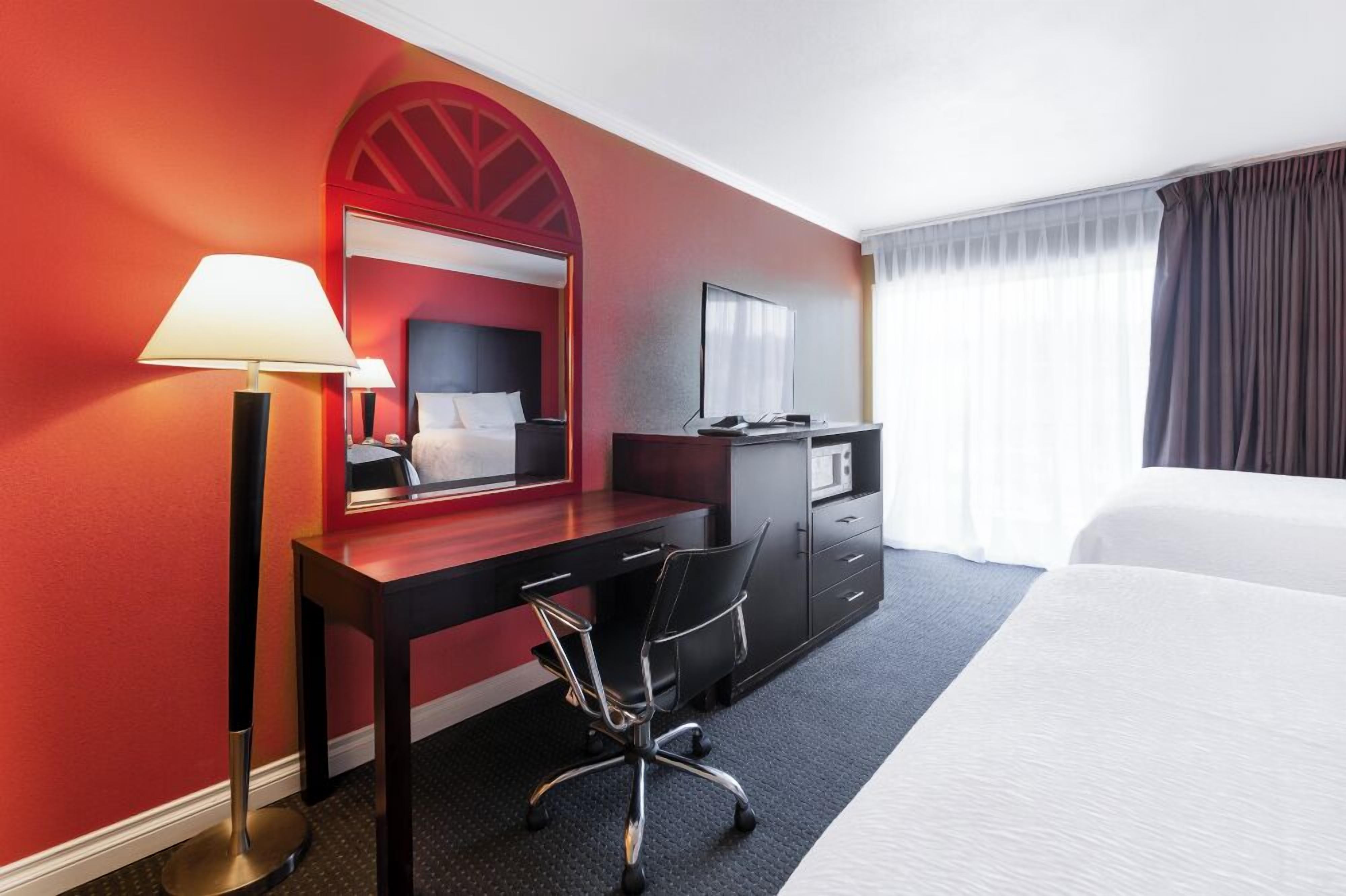 A hotel room with a bed, a desk with a chair, a desk lamp, a dresser, and a large mirror.