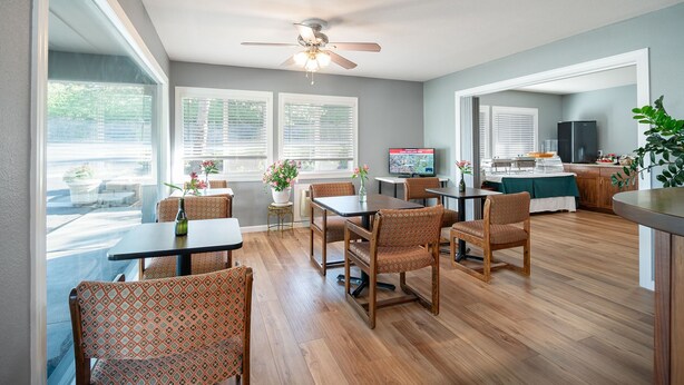 A spacious dining area with wooden tables and chairs, large windows with blinds, a ceiling fan, and a kitchen area with a refrigerator and counter. - Pullman Garden Inn (Pullman)