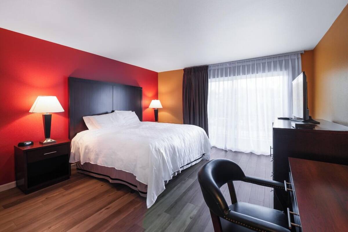 A hotel room with a bed, two bedside lamps, a desk, a chair, and a large window with curtains.