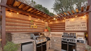 Cocina exterior con pared de piedra, una parrilla, horno empotrado y refrigerador. Hay un cartel que indica información de contacto de emergencia.