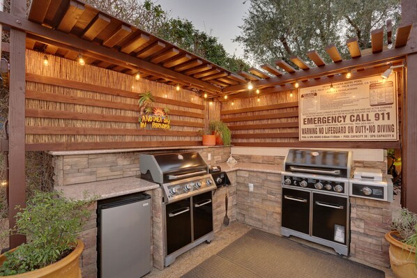 An outdoor kitchen with a stone wall, a grill, a built-in oven, and a refrigerator. There is a sign indicating emergency contact information. - Desert Riviera Hotel (Palm Springs)