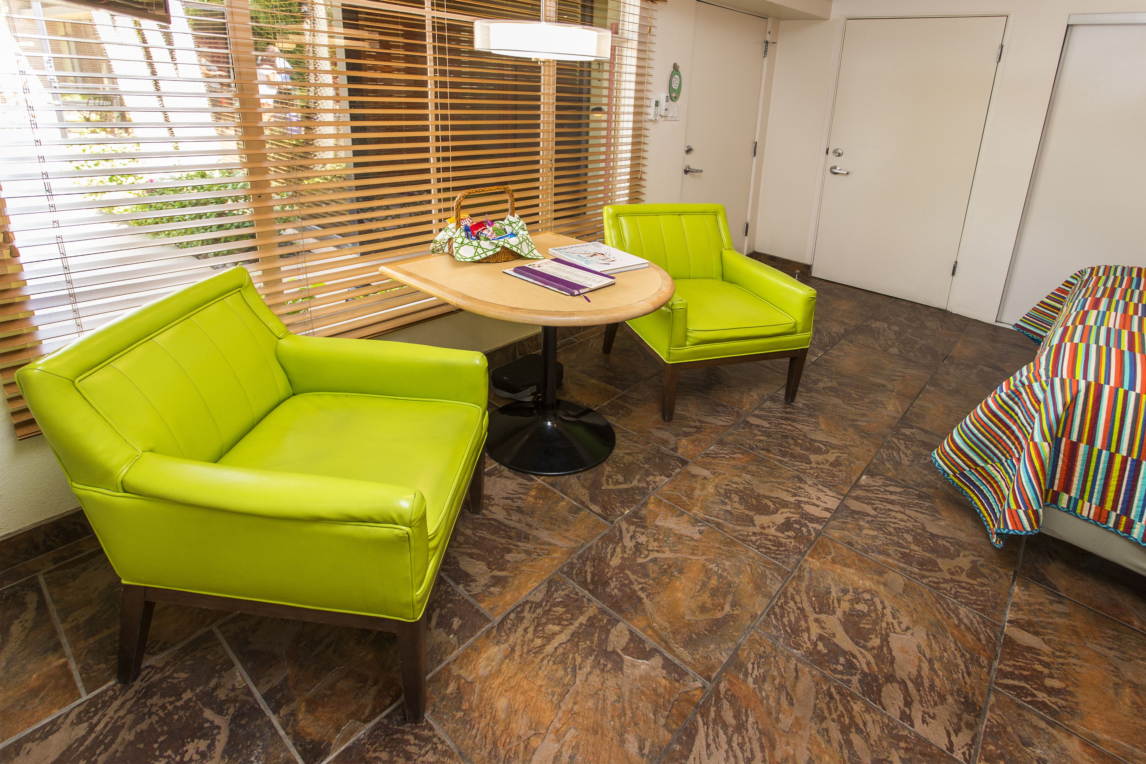 A room with a green sofa, a green armchair, a small round table with a bag and book, and a colorful striped bed.