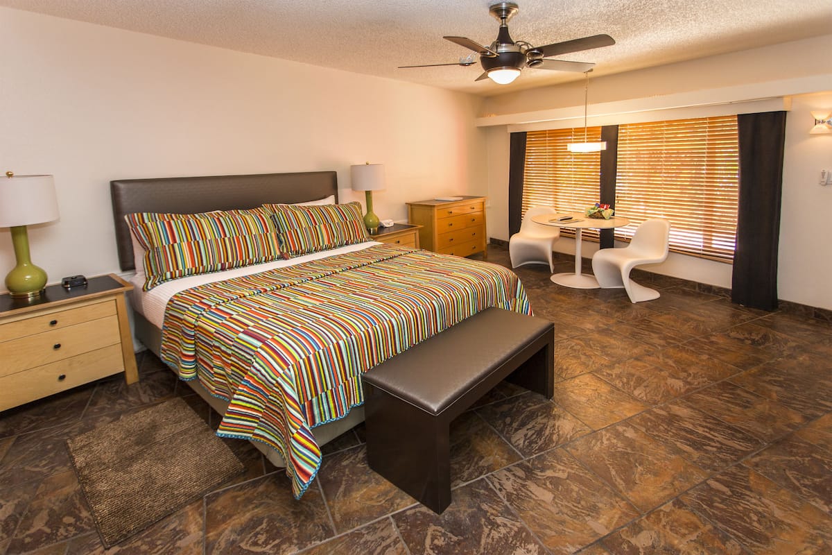 A hotel room with a large bed, a desk with a chair, a ceiling fan, and a window with blinds.