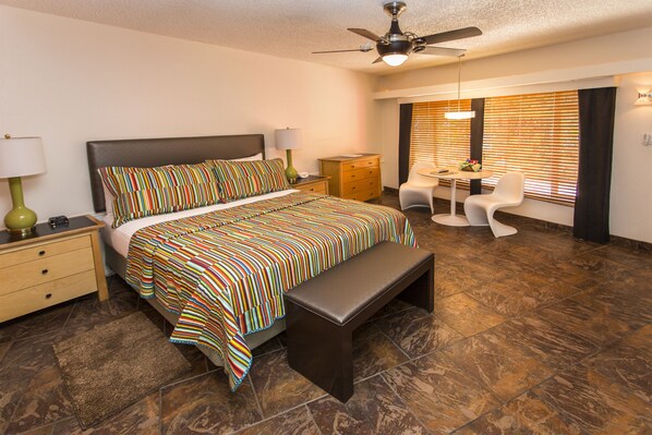 A hotel room with a large bed, a desk with a chair, a ceiling fan, and a window with blinds. - Desert Riviera Hotel (Palm Springs)
