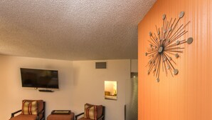 Una sala de estar con televisión, tres sillas, un adorno de pared y una ventana con persianas.