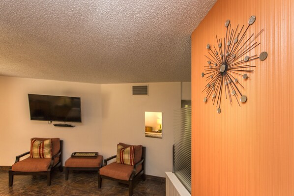 A living room with a television, three chairs, a decorative wall piece, and a window with blinds. - Desert Riviera Hotel (Palm Springs)