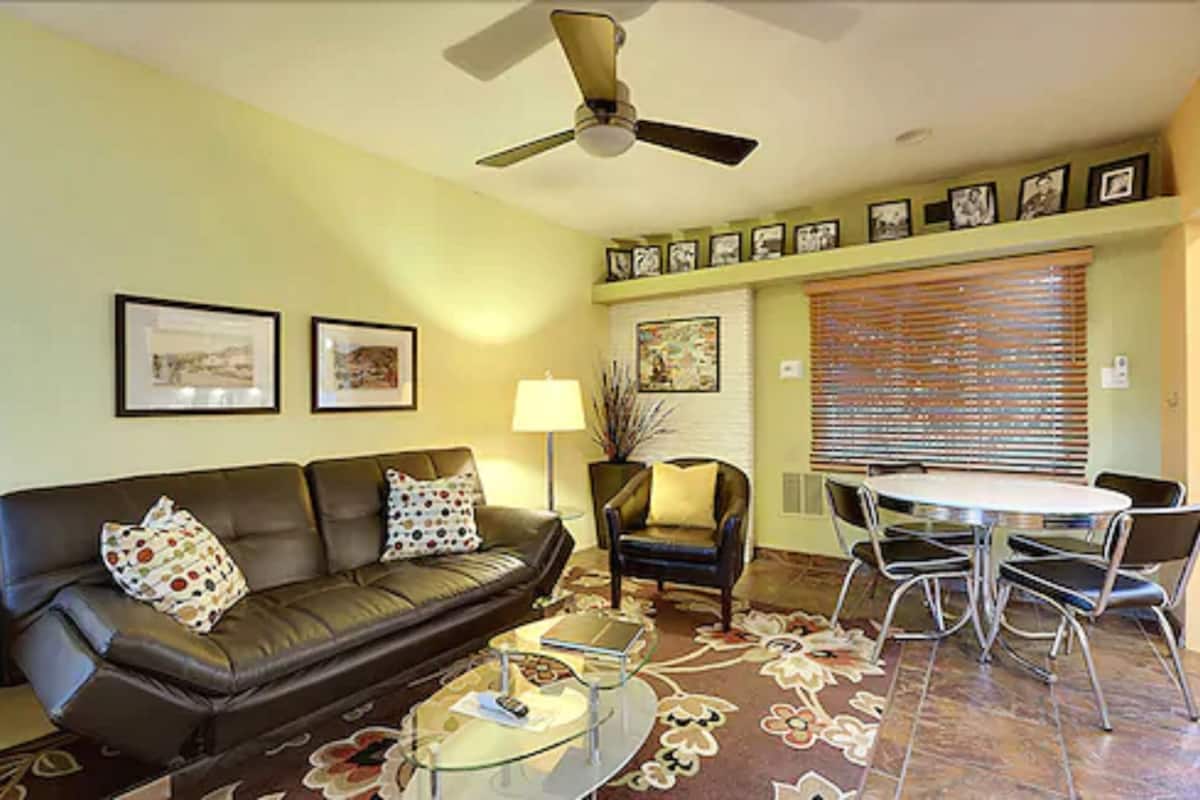 A living room with a brown leather sofa, a glass coffee table, a dining table with chairs, a ceiling fan, and framed pictures on the wall.