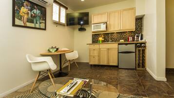 Una cocina compacta con microondas, un lavamanos y una estantería para vinos. Hay una zona de comedor con una mesa y dos sillas. Un televisor está montado en la pared sobre la cocina.