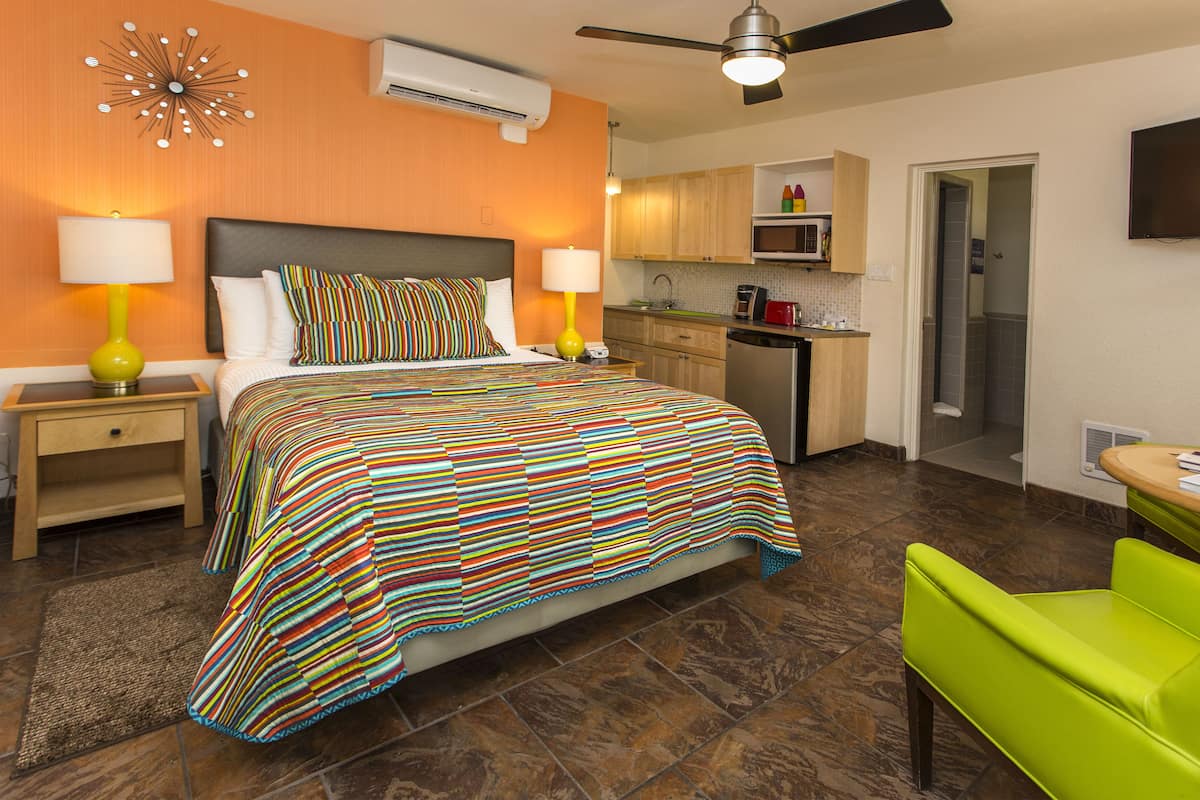 A hotel room with a bed, nightstands, a green chair, a kitchen area, and a TV.