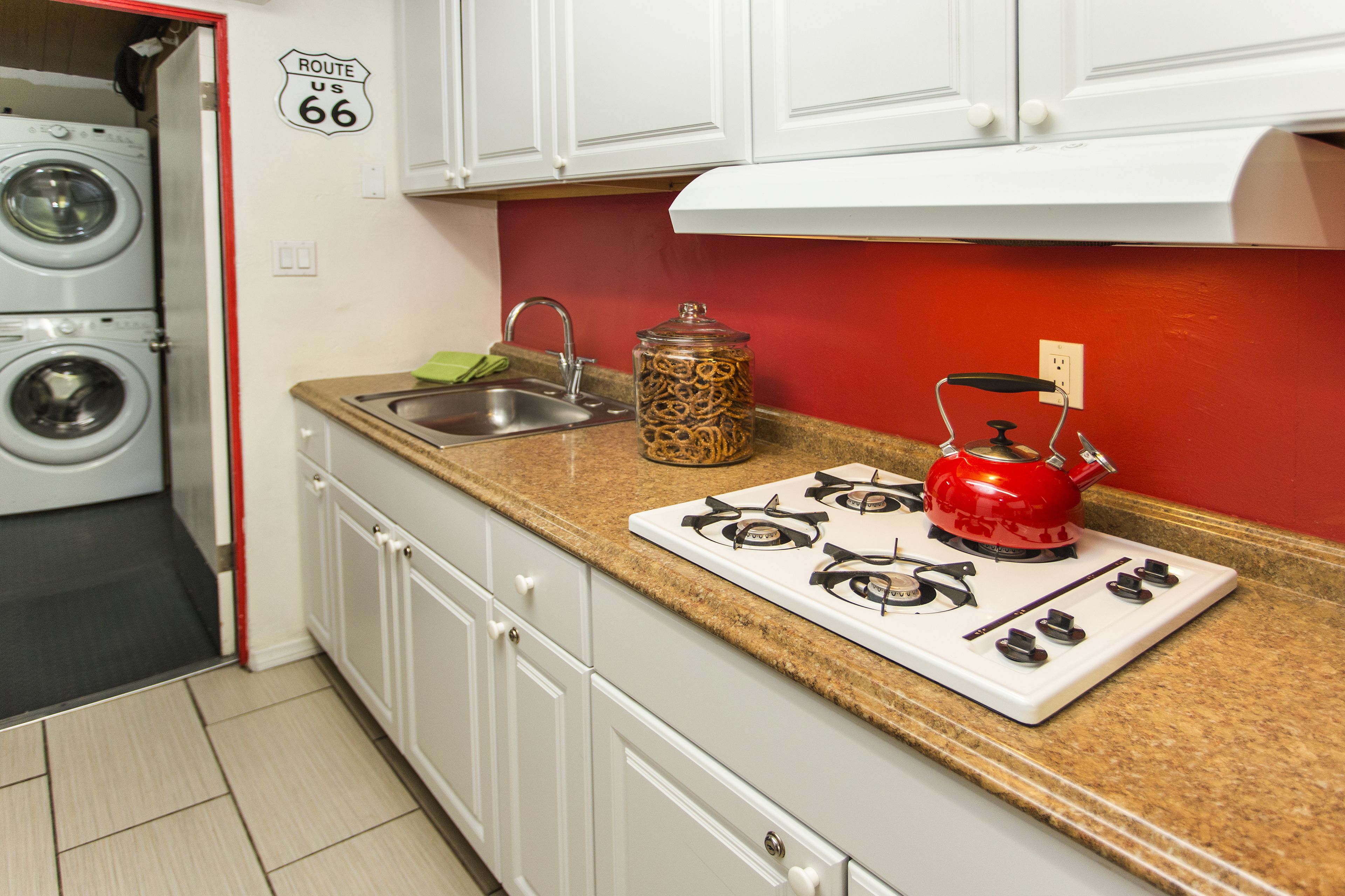 A compact kitchen with a gas stove, a red kettle, a jar of spaghetti, and a washing machine in the background.