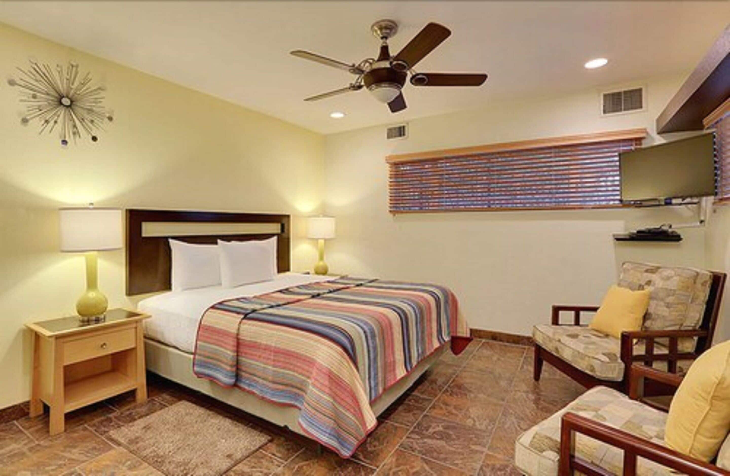 A bedroom with a bed, bedside lamps, a ceiling fan, a window with blinds, and a TV on a stand.