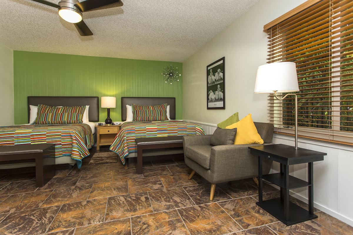 A hotel room with two beds, a bench, a grey armchair, a table, and a window with blinds.