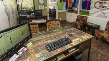 A room with a wooden desk, computer, and various items. There are chairs, a clock, and a wall with hanging items.