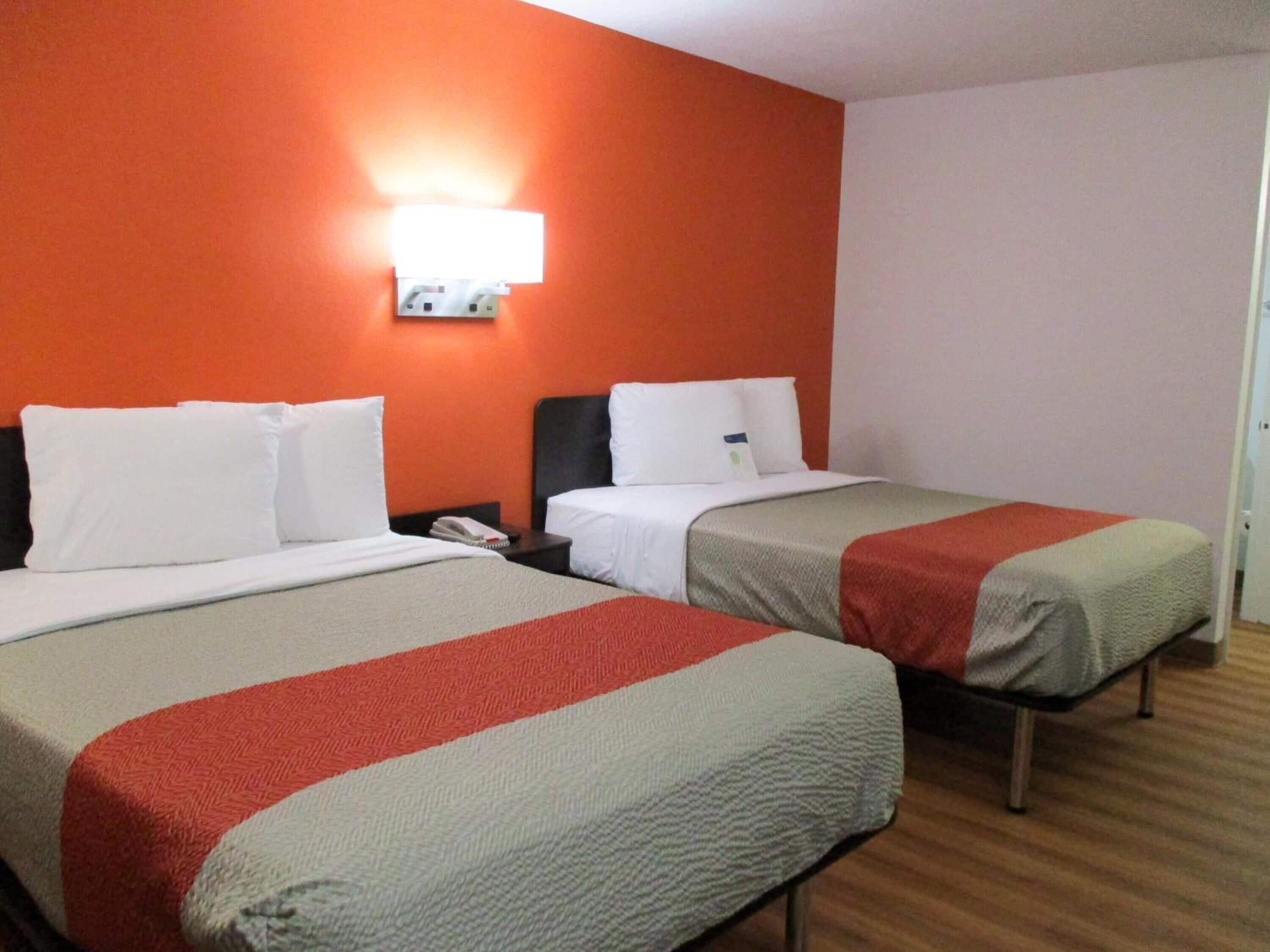 Une chambre d’hôtel avec deux lits, chacun étant équipé de draps blancs et d’une couverture rouge et grise. On y trouve une table de chevet avec un téléphone et une lampe fixée au mur.