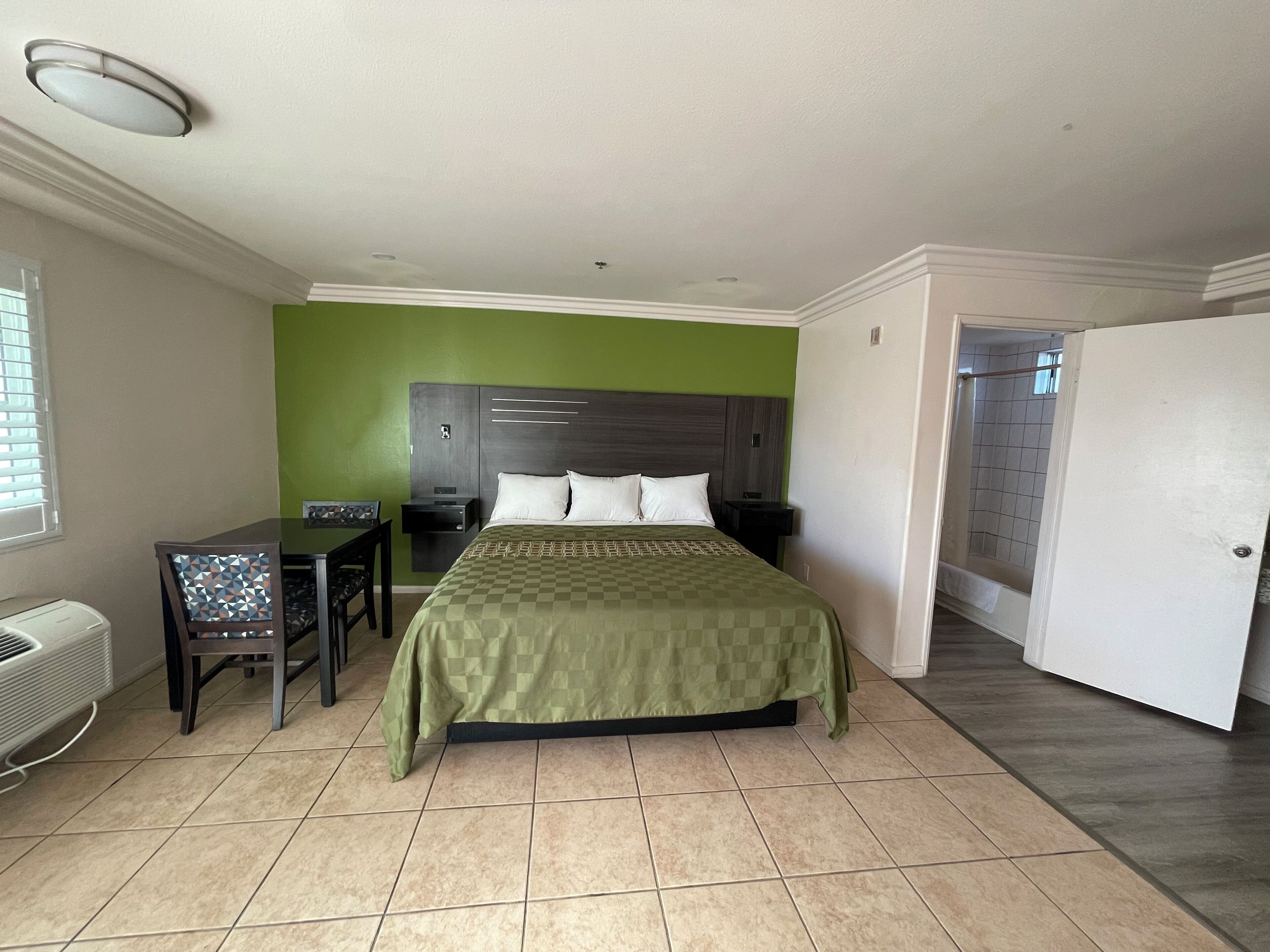 A hotel room with a bed, a desk, and a chair. There is a window with blinds, a door, and a tiled floor.