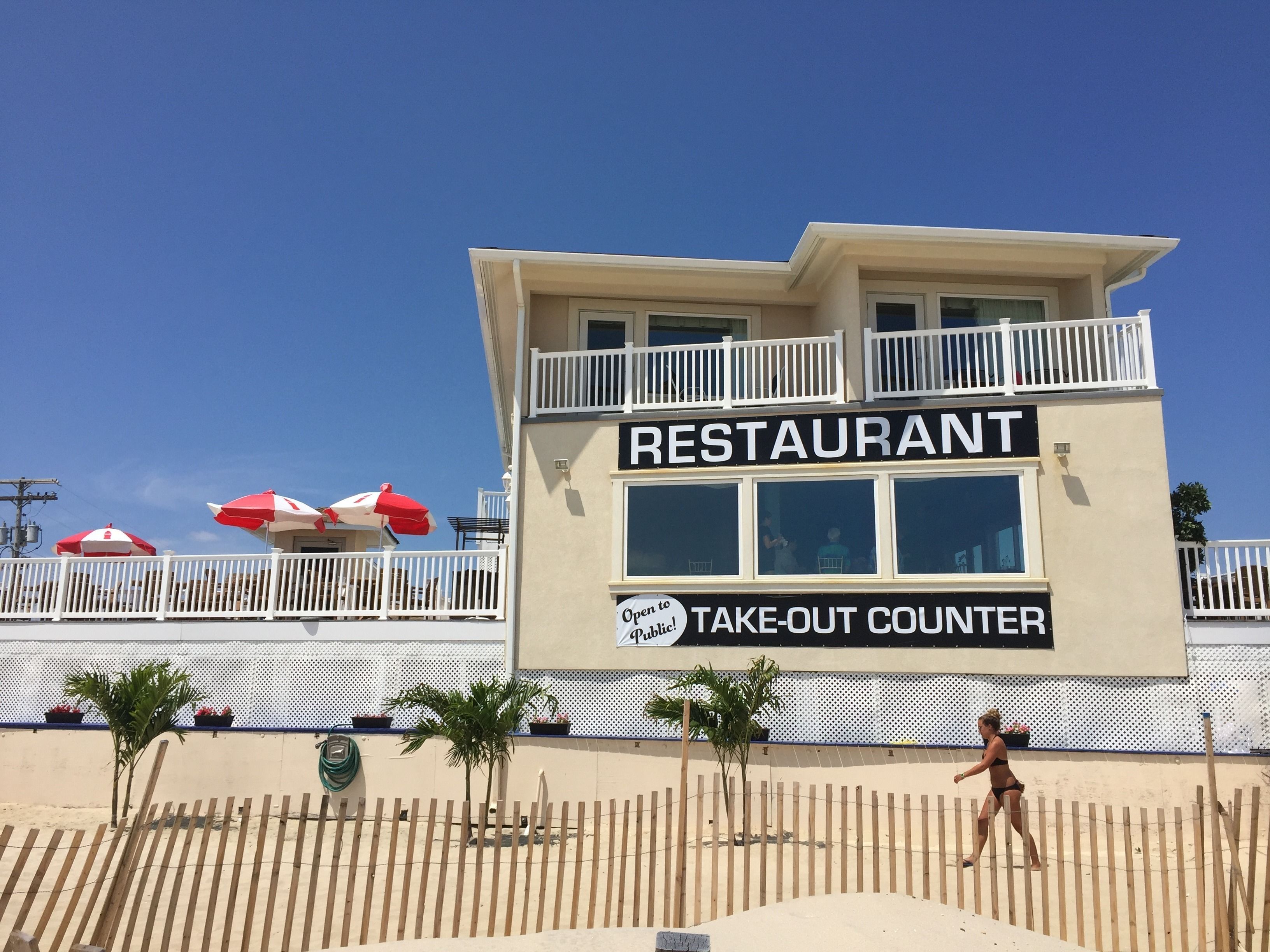 A beachside restaurant with a take-out counter, outdoor seating with umbrellas, and a wooden fence.