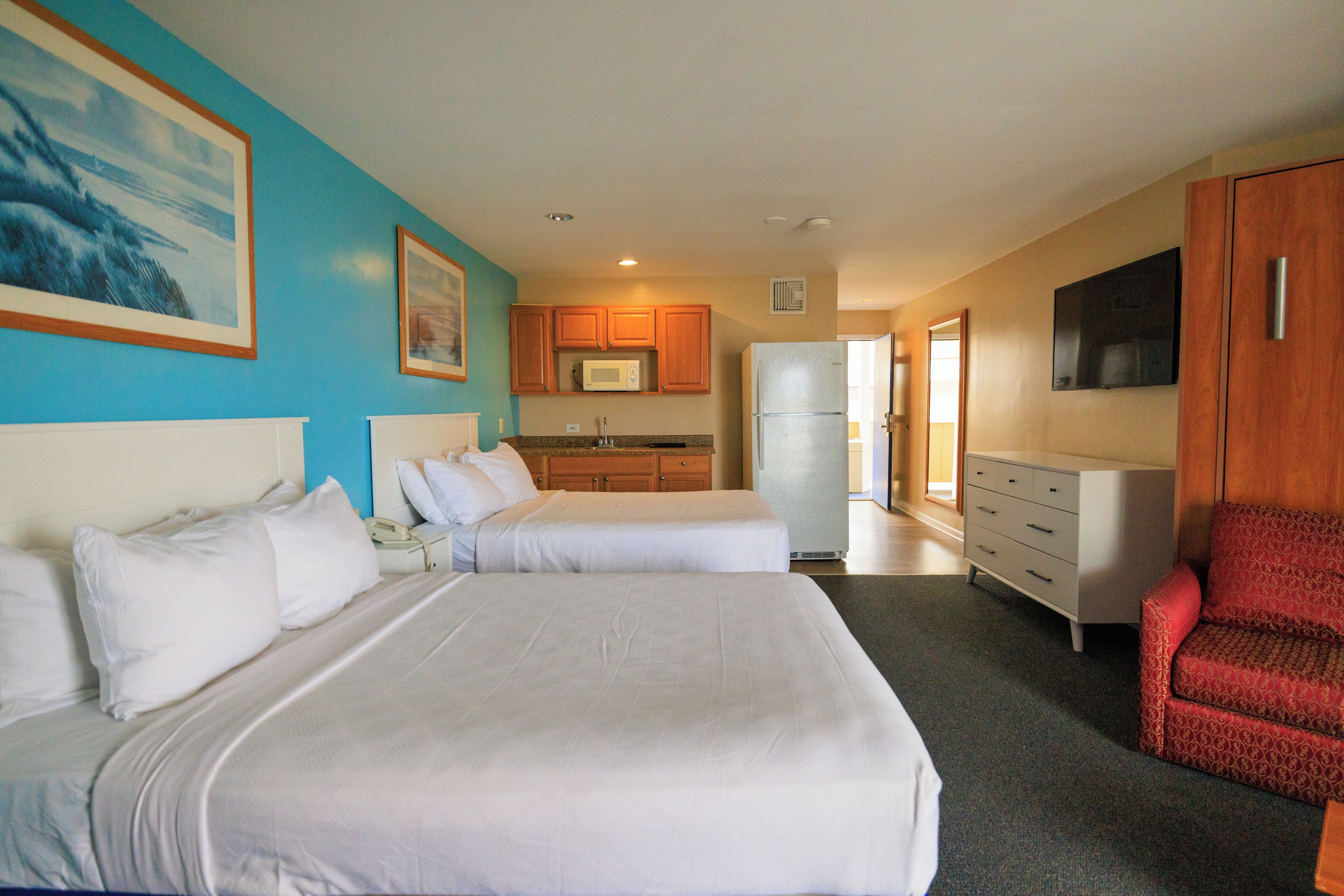 A hotel room with two beds, a kitchenette, a TV, and a red chair.