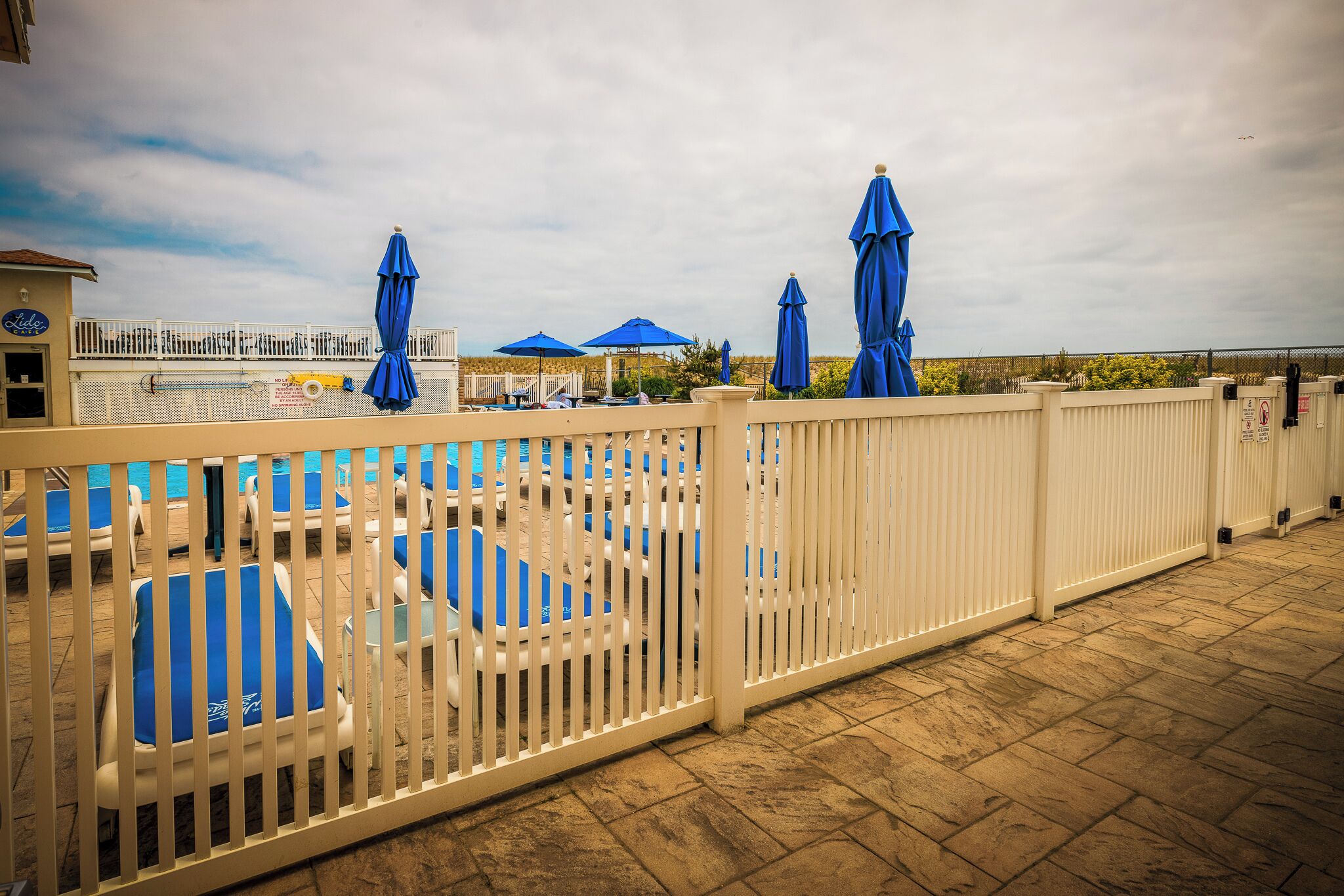 Un área de piscina con sillas y sombrillas azules, una cerca blanca y vistas al cielo.
