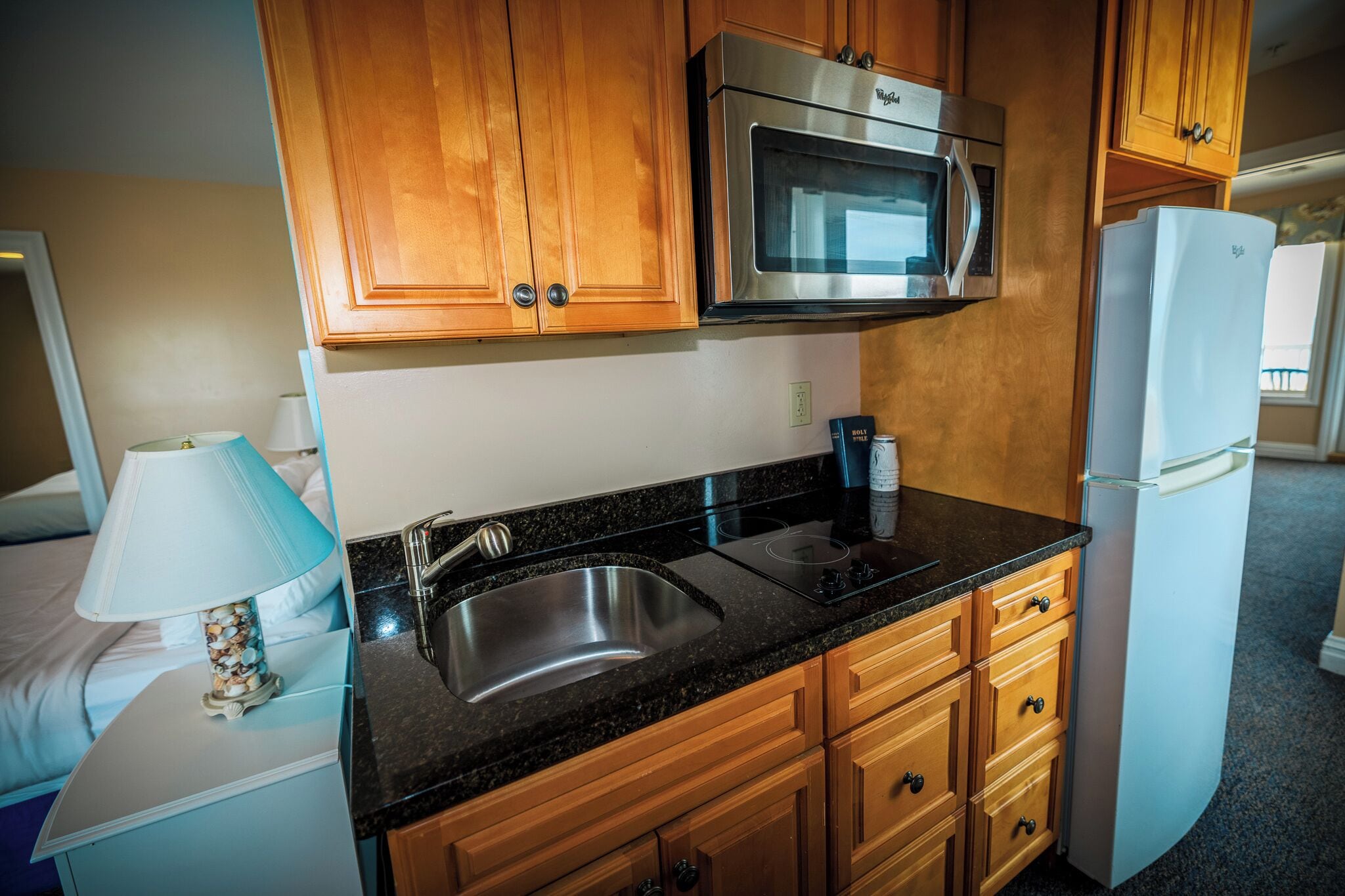 A compact kitchen with wooden cabinets, a stainless steel microwave, a built-in oven, a sink, and a white refrigerator.