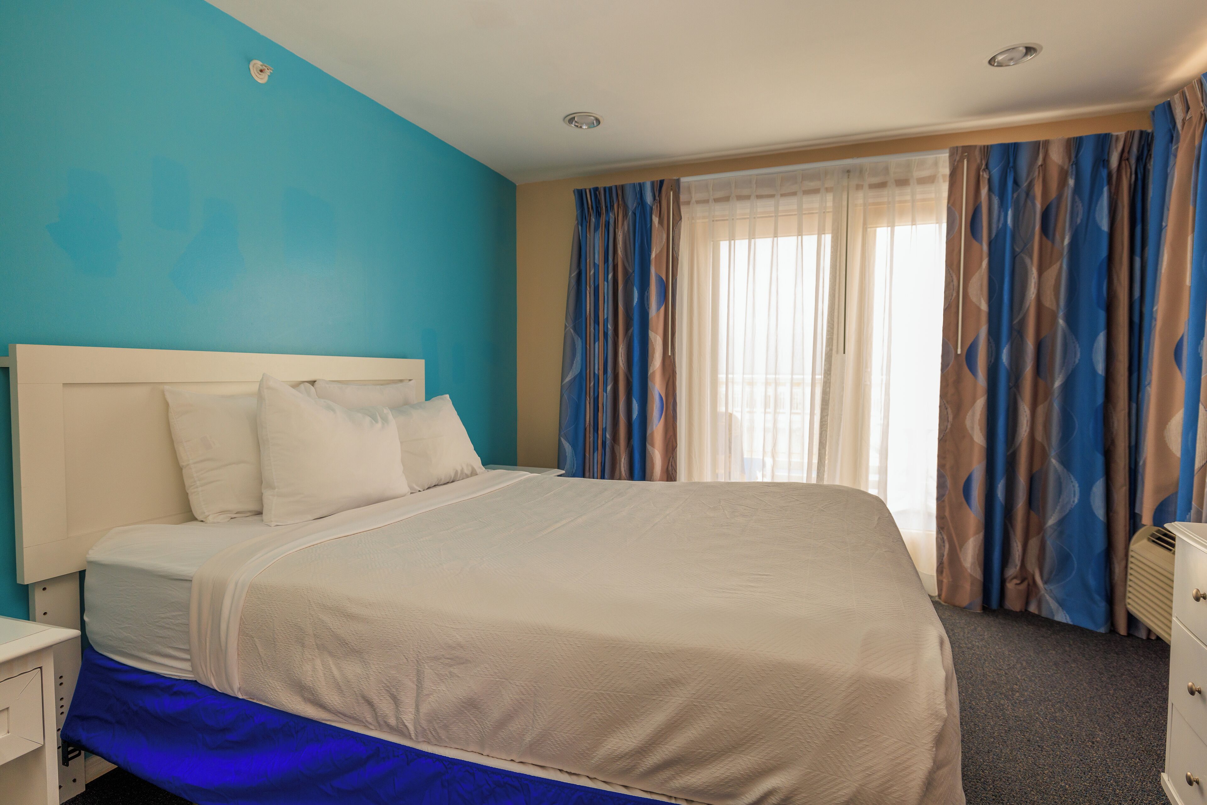 A hotel room with a large bed, a nightstand, a dresser, and a window with curtains.