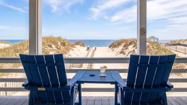 A beach view from a covered area with two blue chairs and a table, a no-smoking sign, and a gazebo in the background. - Blue Heron Motel (Nags Head)